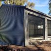 Front view of a gray corrugated metal shed with three large open bays and a concrete floor, under construction in a backyard.
