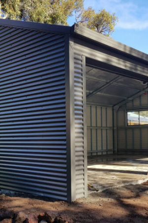 Front view of a gray corrugated metal shed with three large open bays and a concrete floor, under construction in a backyard.