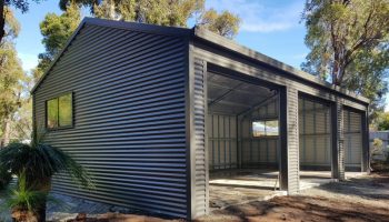 Front view of a gray corrugated metal shed with three large open bays and a concrete floor, under construction in a backyard.