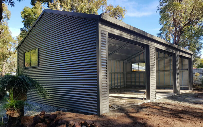 Front view of a gray corrugated metal shed with three large open bays and a concrete floor, under construction in a backyard.