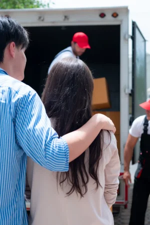 Couple stands by an open moving truck as movers load cardboard boxes inside the trailer.