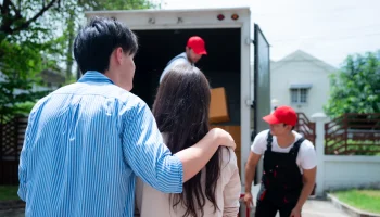 Couple stands by an open moving truck as movers load cardboard boxes inside the trailer.
