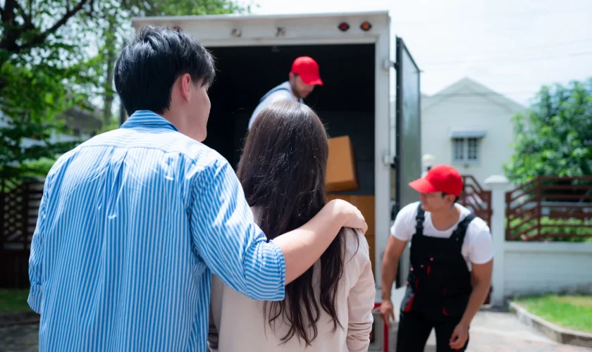 Couple stands by an open moving truck as movers load cardboard boxes inside the trailer.