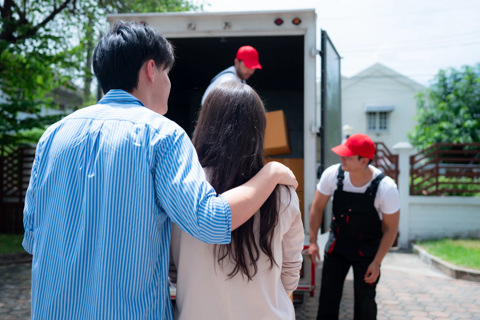 Couple stands by an open moving truck as movers load cardboard boxes inside the trailer.