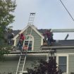 Roofing crew repairing shingles on a gray suburban house; a tall ladder leans against the peak as workers move materials on the roof.