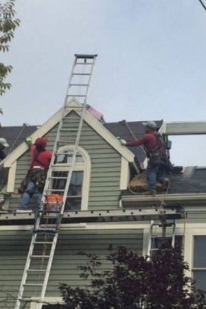 Roofing crew repairing shingles on a gray suburban house; a tall ladder leans against the peak as workers move materials on the roof.