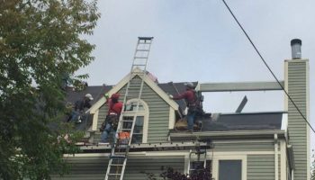 Roofing crew repairing shingles on a gray suburban house; a tall ladder leans against the peak as workers move materials on the roof.