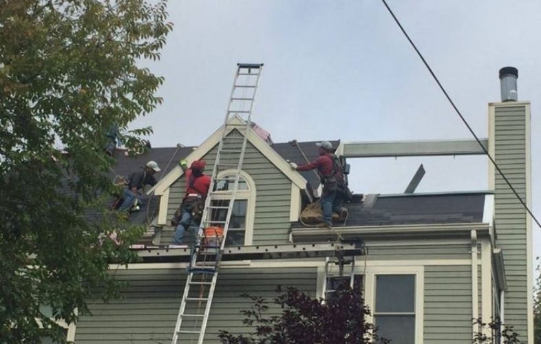 Roofing crew repairing shingles on a gray suburban house; a tall ladder leans against the peak as workers move materials on the roof.