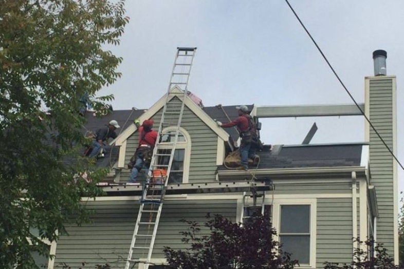 Roofing crew repairing shingles on a gray suburban house; a tall ladder leans against the peak as workers move materials on the roof.